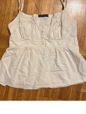 Vit brandy topp med knappar - Söt vit linnetopp från Brandy melville med smala axelband och dekorativa knappar framtill. Toppen har en lätt rynkad byst och luftig passform, perfekt för varma dagar. Enkel och stilren design som passar till mycket. Passar en xs/s