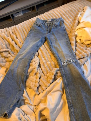 Ljusblå slitna jeans från Gina Tricot - Säljer ett par ljusblå jeans från Gina Tricot med boot cut och slitning vid ena knät. Klassisk femficksmodell med dragkedja och knapp. Jeansen är i denim och har en avslappnad, trendig look.