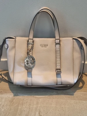 Beige handväska från Guess - Snygg beige handväska från Guess med silverfärgade detaljer och dekorativa nitar på handtagen. Väskan har både handtag och axelrem samt en stor Guess-logga i metall som hänger framtill. Tillverkad i konstläder med strukturerad yta.