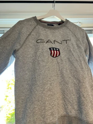 Grå sweatshirt från GANT - Snygg grå sweatshirt från GANT med broderad logga och amerikanskt inspirerat sköldmärke på bröstet. Tröjan har rund halsringning och långa ärmar. Tillverkad i mjukt sweatshirtmaterial som passar perfekt till en avslappnad stil. Står 170, passar fortfarande mig och är 177! 