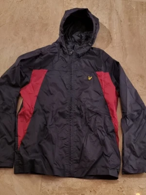 Svart och röd vindjacka med huva - Snygg vindjacka från Lyle & Scott i svart med röda paneler på sidorna. Jackan har huva, hel dragkedja framtill och den klassiska loggan på bröstet. Tillverkad i lätt, vindtätt material som passar perfekt för blåsiga dagar. Stl L