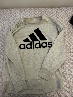 Grå sweatshirt från Adidas - Snygg grå sweatshirt från Adidas med stort svart logotryck på bröstet. Tröjan har rund halsringning, långa ärmar och breda muddar vid ärmslut och nederkant. Perfekt för en avslappnad och sportig stil.