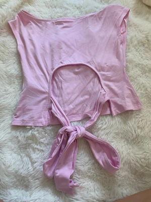 Ljusrosa topp med knyt och öppen detalj - Trendig ljusrosa topp i mjuk bomull med öppen detalj framtill och breda knytband i midjan. Toppen är ärmlös och har en lätt croppad passform som ger en modern look. Perfekt att styla med höga jeans eller kjol för en cool vibe.