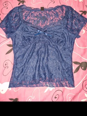 Blå spetsblus med rosett - Blå blus i spets med korta ärmar och v-ringad urringning. Toppen har en liten rosett framtill och är transparent med blommigt spetsmönster. Perfekt för dig som gillar romantiska och feminina detaljer.