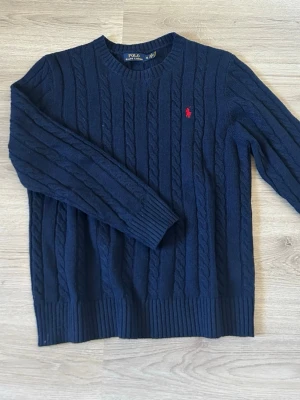 Mörkblå kabelstickad tröja Polo Ralph Lauren - Kabelstickad mörkblå tröja från Polo Ralph Lauren med rund halsringning och ribbade muddar. Tröjan har det klassiska röda Polo-logot broderat på bröstet och är tillverkad i mjukt material. Perfekt för dig som gillar stilren och tidlös design.