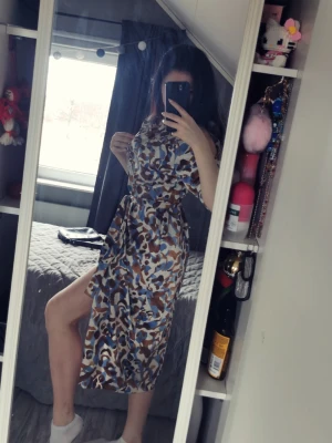 Mönstrad midiklänning med knappar och bälte - Säljer en snygg midiklänning med blått, brunt och vitt abstrakt mönster. Klänningen har korta ärmar, markerad midja med knytskärp och en hög slits framtil (kan kanppa ihop). Luftigt material och avslappnad passform, perfekt för dig som gillar trendiga statement-plagg.