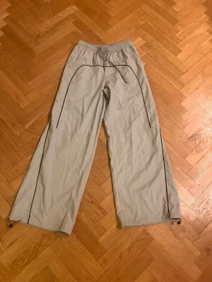 Vida ljusgrå mjukisbyxor med svarta detaljer - Säljer ett par ljusgrå mjukisbyxor med vida ben och svarta kontrastsömmar. Byxorna har resår i midjan och justerbara snören vid bensluten. Perfekta för en avslappnad och trendig look. Materialet känns lätt och bekvämt.Skriv gärna för mått! 💕