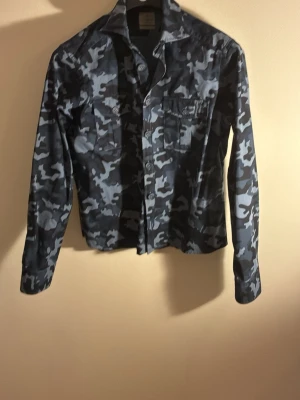 Mörkblå camouflage skjorta - Snygg långärmad skjorta i mörkblått camouflage-mönster. Skjortan har två bröstfickor med lock och knappar framtill. Perfekt för dig som vill sticka ut med en cool och trendig stil.