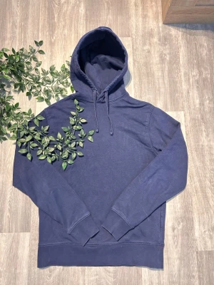 Mörkblå basic hoodie med huva - En stilren mörkblå hoodie med huva. Hoodien har långa ärmar, ribbade muddar vid ärmslut och nederkant samt en klassisk passform. Perfekt för en avslappnad look och enkel att matcha med andra plagg. Materialet känns mjukt och bekvämt.