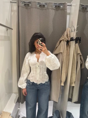 Vit spetsblus med puffärm - Romantisk vit blus i spets med v-ringning och knappar framtill. Blusen har långa puffärmar och figurnära passform med vågig kant nedtill. Perfekt för dig som gillar en feminin och trendig stil.