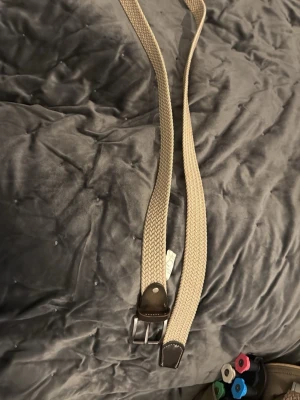 Beige flätat bälte med skinn-detaljer - Snyggt beige flätat bälte med bruna skinn-detaljer vid spännet. Bältet har ett klassiskt silverfärgat metallspänne och passar perfekt till jeans eller chinos. Enkel och stilren design som funkar till många outfits.