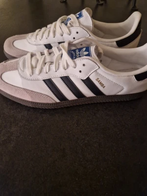 Adidas Samba vita och svarta sneakers - Klassiska Adidas Samba sneakers i vitt läder med svarta ränder och detaljer. Tåpartiet är i ljusgrå mocka och yttersulan är brun. Skorna har blå Adidas-logga på plösen och texten 'SAMBA' i guld på sidan. Snygg retrostil som alltid funkar.