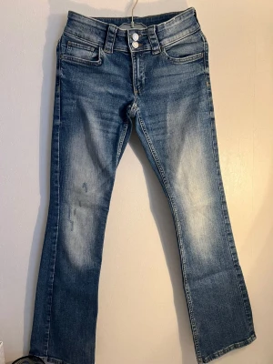 Blå bootcut jeans med dubbla knappar - Snygga blå bootcut jeans med dubbla knappar i midjan och slitna detaljer framtill. Jeansen har klassiska fem fickor, dekorativa bakfickor med lock och vita sömmar. Perfekta för en avslappnad och trendig look.