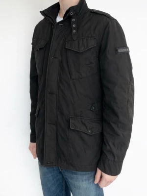 Svart Field Jacka från Woolrich - Snygg svart parkajacka från Woolrich med fyra fickor framtill, hög krage med tryckknappar och axelklaffar. Jackan har diskret logga på ärmen och stängs med dragkedja och knappar. Perfekt för kyligare dagar.