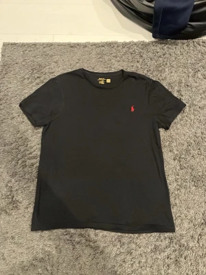 Svart t-shirt från Ralph Lauren - Svart t-shirt från Ralph Lauren med klassisk röd broderad logga på bröstet. Enkel och stilren design med rund hals och korta ärmar. Perfekt till jeans eller shorts för en clean look.