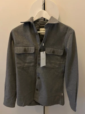 Grå overshirt med fickor - Stilren grå overshirt i mjukt tyg med klassisk krage och två stora bröstfickor med lock och knappar. Knäppning framtill med matchande knappar. Perfekt lager-på-lager-plagg för en avslappnad look.