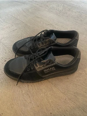 Svarta Versace Jeans Couture sneakers - Snygga svarta sneakers från Versace Jeans Couture med läder- och mockadetaljer. Skorna har perforerad tå, svarta skosnören och tydlig logga på sidan samt på sulan. Perfekta för dig som gillar stilrena och exklusiva sneakers.