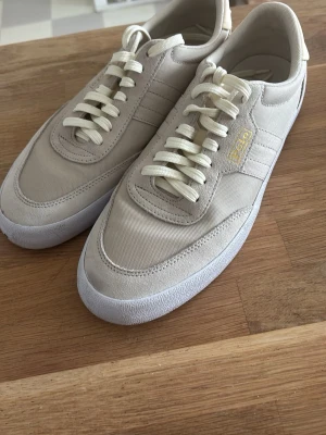 Beige Polo Ralph lauren sneakers - Snygga beige polo Ralph lauren  sneakers och guldfärgad logga. Ovandelen är i mocka och textil, med vita snören och vit sula. Perfekta för en clean och stilren look.