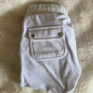 Vita mjukisbyxor från Juicy Couture - Säljer ett par vita mjukisbyxor från Juicy Couture med klassisk bakficka med lock och silverfärgade knappar. Byxorna har dragsko i midjan och en broderad JC-logga på ena fickan. Perfekta för en chill och avslappnad stil.