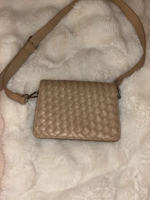 Beige väska - Snygg beige axelväska med bred justerbar rem och flätad design på framsidan. Väskan har en stilren, rektangulär form och är gjord i ett glansigt konstlädermaterial. Perfekt för dig som gillar trendiga och minimalistiska accessoarer. Vafan är från NA-KD som man kan se på bild 3. Pris kan diskuteras. 