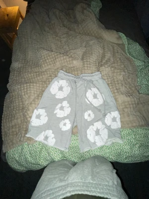 Grå shorts med vita blommor - Säljer ett par grå shorts med vita blommönster och resårmidja med snörning. De har en avslappnad passform och är perfekta för sommaren. Materialet känns mjukt och bekvämt, troligen bomull eller en bomullsblandning.