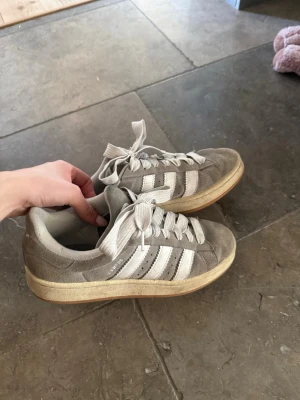 Beige Adidas sneakers i mocka - Snygga gråa Adidas sneakers i mocka med klassiska vita ränder på sidorna och vit sula med gummidetaljer. Skorna har låga skaft och breda vita skosnören. Perfekta för en avslappnad streetstyle-look.