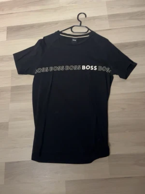 Hugo Boss T-shirt - Snygg svart t-shirt från BOSS med vit logga tryckt flera gånger över bröstet. Klassisk rund halsringning och korta ärmar. Perfekt för dig som gillar stilrena plagg med tydlig branding.