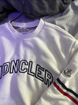 Vit  sweatshirt med logga - Säljer en vit sweatshirt från Moncler med stor broderad logga över bröstet och klassisk Moncler-patch på ärmen. Tröjan har rund halsringning och ribbade muddar. Ärmen har snygga ränder i rött och blått för extra detalj.