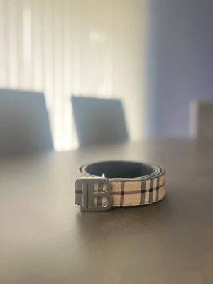 Burberry bälte med rutigt mönster - Snyggt bälte från Burberry med klassiskt rutigt mönster i beige, rött och svart. Bältet har ett stilrent metallspänne med Burberrys logotyp. Perfekt accessoar för att lyfta din outfit.