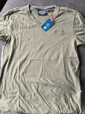 Olivgrön t-shirt från Superdry (oanvänd) - Olivgrön t-shirt från Superdry Athletics med v-ringning och broderad logga på bröstet. Tillverkad i mjuk bomull och har en klassisk passform med korta ärmar. Perfekt för en avslappnad och stilren look.