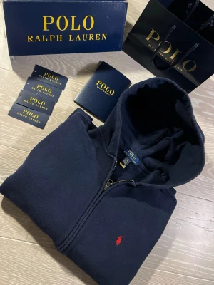 Mörkblå hoodie från Polo Ralph Lauren - Snygg mörkblå hoodie från Polo Ralph Lauren med dragkedja och klassisk röd broderad logga på bröstet. Hoodien har huva och är tillverkad i mjukt material som känns skönt mot huden. Perfekt för en avslappnad och stilren look.