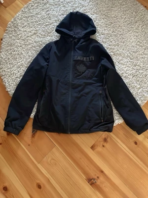 Svart vindjacka från Zavetti - Snygg svart vindjacka från Zavetti med huva och dragkedja framtill. Jackan har en diskret logga och lönnlöv på bröstet samt två fickor med dragkedja. Materialet är slätt och vattenavvisande, perfekt för blåsiga dagar.