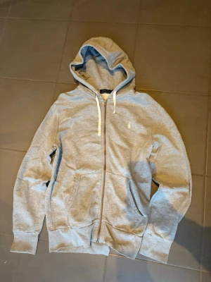 Grå zip-hoodie från Polo Ralph Lauren - Snygg grå hoodie med dragkedja från Polo Ralph Lauren. Snygg design med två fickor fram och broderad logga på bröstet. Mjuk bomullsblandning och ribbade muddar för extra komfort. Den är i nyskick och jag har använt den ett fåtal gånger. Säljer den då jag inte använder den längre😊