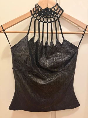Svart glittrig halterneck topp - Unik svart glittrig topp med halterneck och smala axelband. Toppen har ett coolt rutnät vid halsen med små stenar och ett lätt skimrande material med ormskinnsinspirerat mönster. Perfekt för dig som vill sticka ut.