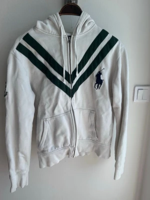 Vit hoodie Polo Ralph Lauren Wimbledon - Vit vintage hoodie från Polo Ralph Lauren med gröna V-formade ränder över bröstet och Wimbledon-märke på ärmen. Dragkedja framtill, två fickor och klassisk huva. Stort Polo-logotyp på bröstet. Perfekt för dig som gillar sportig och stilren look.