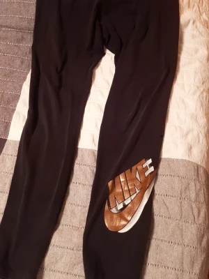 Svarta Nike leggings med metallic-logga - Säljer ett par svarta leggings från Nike med en stor metallicfärgad logga på benet. Materialet är stretchigt och bekvämt, perfekt för att chilla eller träna. Loggan har en cool kopparfärgad finish som verkligen sticker ut.