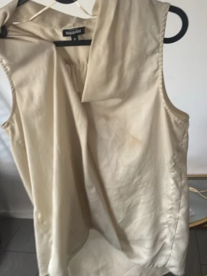Beige ärmlös topp från Kappahl - Stilren beige ärmlös topp från Kappahl med lös passform och unik krage. Tillverkad i ett mjukt, lätt material som ger ett snyggt fall. Perfekt att matcha med jeans eller kjol för en trendig look.