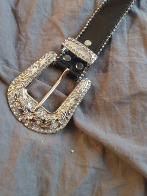 Svart bälte med glittrande spänne - Snyggt svart bälte med ett stort, glittrande silverspänne dekorerat med stenar och detaljerade mönster. Bältet har silverfärgade nitar längs kanterna och ger en riktigt statement look till outfiten.