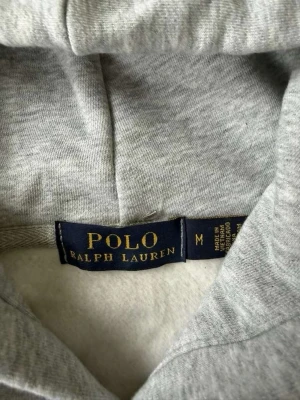 Grå sweatshirt Polo Ralph Lauren M - Snygg grå sweatshirt från Polo Ralph Lauren med tryck av tre polospelare i blått, rött och svart framtill. Tröjan har rund halsringning och mjukt material, perfekt för en avslappnad och sportig stil.