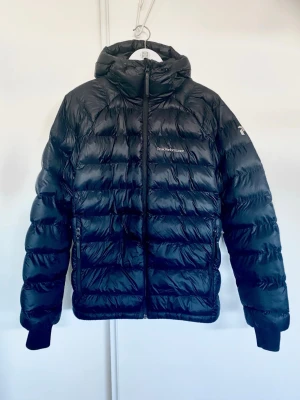 Svart dunjacka från Peak Performance - Snygg svart dunjacka från Peak Performance med huva och dragkedja framtill. Jackan har quiltad design, två sidofickor med dragkedja och diskret logga på bröstet. Materialet är matt och ger en modern look. Använd 2 gånger, men fick ett hål på ryggen samt i fickan. Hör av er vid funderingar.