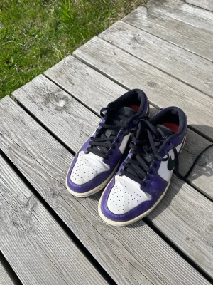 Nike Air Jordan 1 Low Court Purple - Säljer ett par Nike Air Jordan 1 Low Court Purple med svart swoosh, vita paneler och lila detaljer. Skorna har svarta skosnören och perforerad tåbox. Klassisk low-top siluett och bekväm passform. Perfekta för dig som gillar streetwear och sneakerskultur.