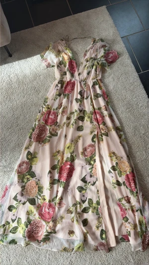 Nelly Eve + Petra Tungården Blommig långklänning med volangärm - Romantisk långklänning med blommigt mönster i rosa, grönt och beige. Klänningen har v-ringning, volangärmar och en luftig, flowy passform. Perfekt för sommarens alla tillfällen. Tillverkad i ett lätt, transparent material med innerfoder.