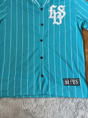 Turkos randig skjorta från FB Sister - Turkos skjorta från FB Sister med vita vertikala ränder, svarta knappar och broderad logga på bröstet. Skjortan har sportig basebollkrage och patch med '92 FBS' nedtill. Perfekt för en avslappnad streetstil.