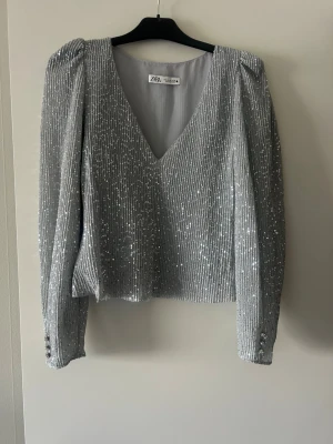Glittrig silverblus från Zara - Säljer en snygg silverfärgad blus från Zara med v-ringning och långa ärmar. Blusen är täckt av glittriga paljetter och har axelvaddar som ger en cool siluett. Knappdetaljer vid ärmslut och rak passform. Perfekt för dig som gillar att sticka ut!