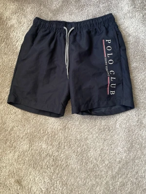 Svarta badbyxor Polo Club - Snygga svarta badbyxor från Polo Club med vit dragsko i midjan och tryckt logga längs ena benet. Klassisk modell med elastisk midja och sportig känsla. Perfekta för stranden eller poolen. Säljer för att jag vuxit ur dem tidigt tyvärr. Använda 5-6 gånger.