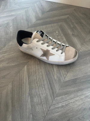 Skor med stjärna  - Snygga vita sneakers från Golden Goose med grå mockadetaljer på tån och en ikonisk beige stjärna på sidan. Skon har grå skosnören och svart hälparti. Perfekt för dig som gillar en trendig och avslappnad stil. Har aldrig använt för dom va för små. dom kommer i en vit box. Priset går att diskutera vid snabb affär 