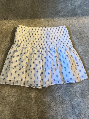 Vit och blå Gina Tricot kjol - Söt vit kortkjol från Gina Tricot med blått grafiskt mönster. Kjolen har smock i midjan och volangdetaljer som ger en luftig och somrig känsla. Storlek 158/164