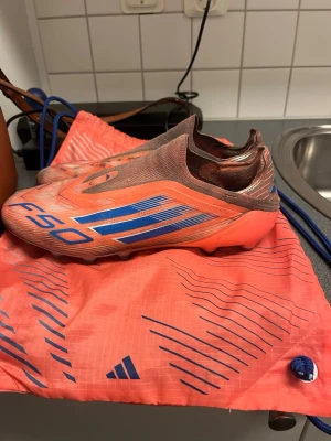 Adidas F50 Laceless elite Ag - Säljer ett par Adidas F50 laceless fotbollsskor i en snygg orange färg med blå detaljer och logga.  Lite slitna fram till på vänster sko. Påse får du med! 