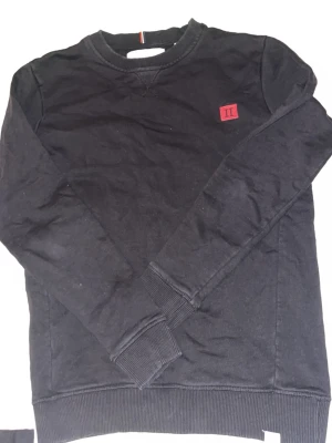 Svart sweatshirt från Les Deux - Svart sweatshirt med rund hals från Les Deux, Tröjan har ribbade muddar vid ärmslut och nederkant samt en röd patch med logga på bröstet. Klassisk och enkel design som passar till mycket. Materialet känns mjukt och bekvämt.