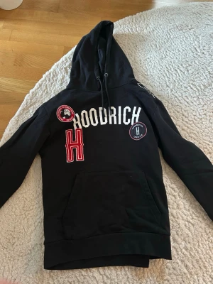 Svart Hoodrich hoodie med patches - Svart hoodie från Hoodrich med stor huva och dragsko. Framsidan har vita och röda patches samt broderade detaljer och loggor. Stor magficka framtill. Tröjan är som helt ny och har bara hängt i min garderob. Inga skador eller så med tröjan. Storlek M